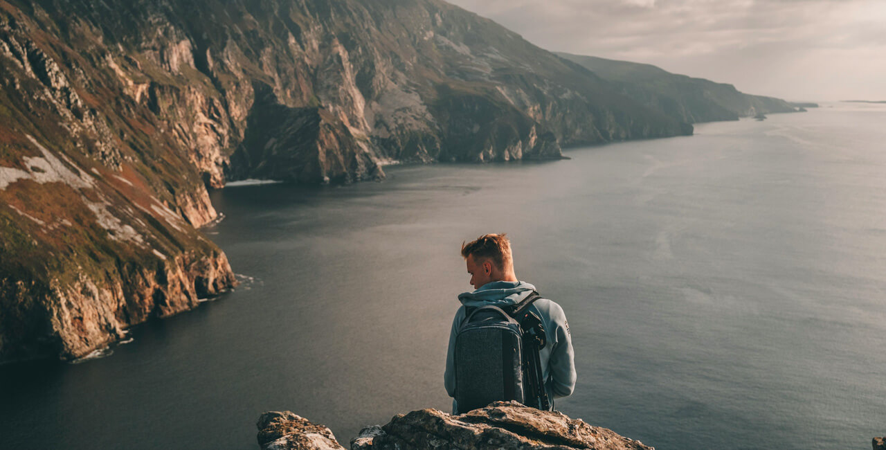 Image of a man sitting on the edge of Sliabh League cliffs, overlooking the vast blue ocean. He is wearing a backpack and admiring the breathtaking scenery.